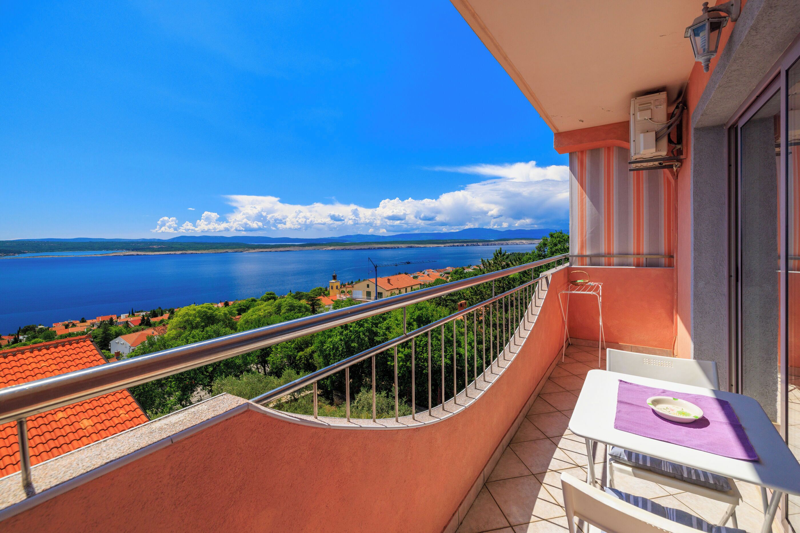 A balcony with a view of a body of water, a railing, a table with a chair, and a window.