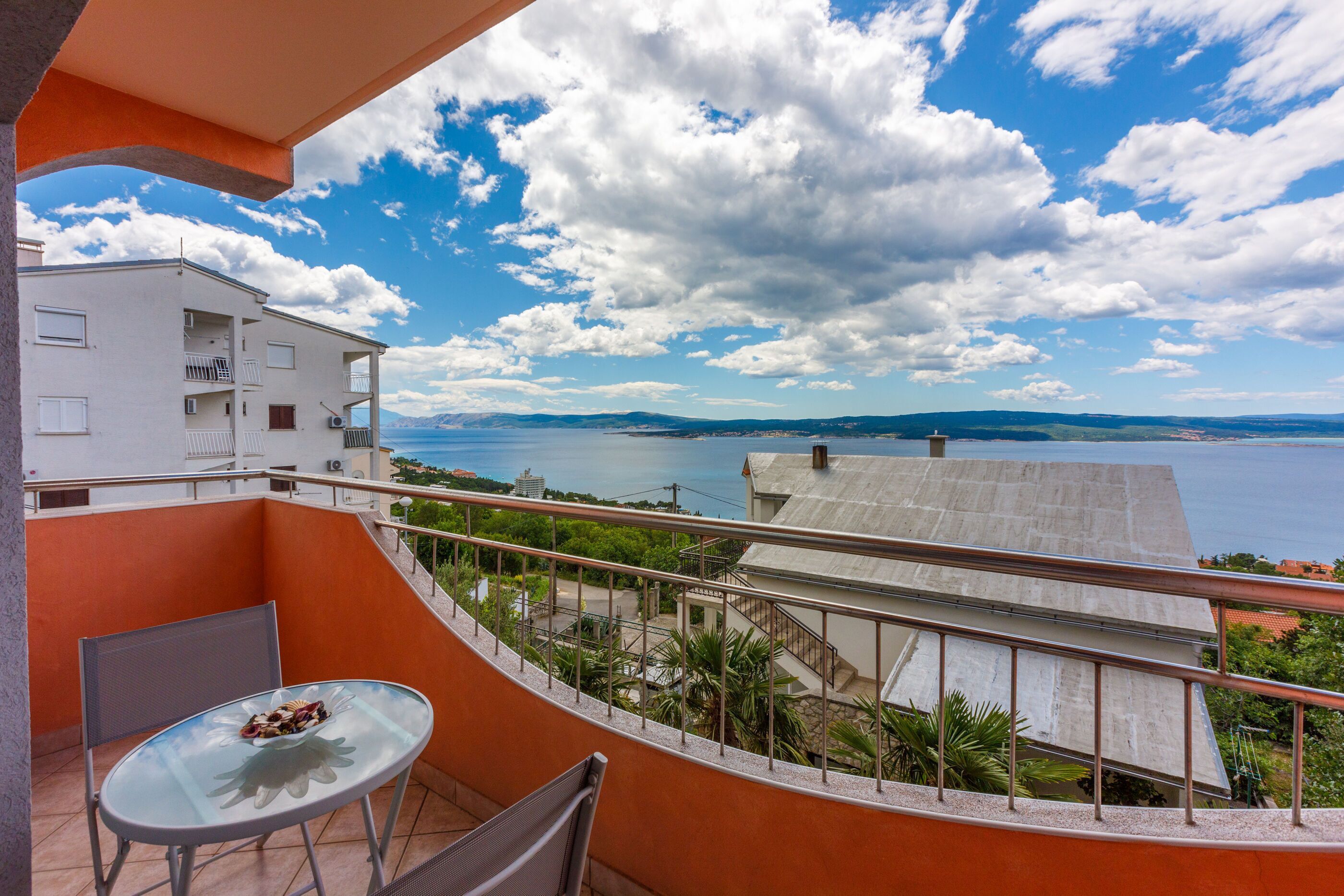 A balcony with a glass table and chairs, overlooking a body of water and distant hills.
