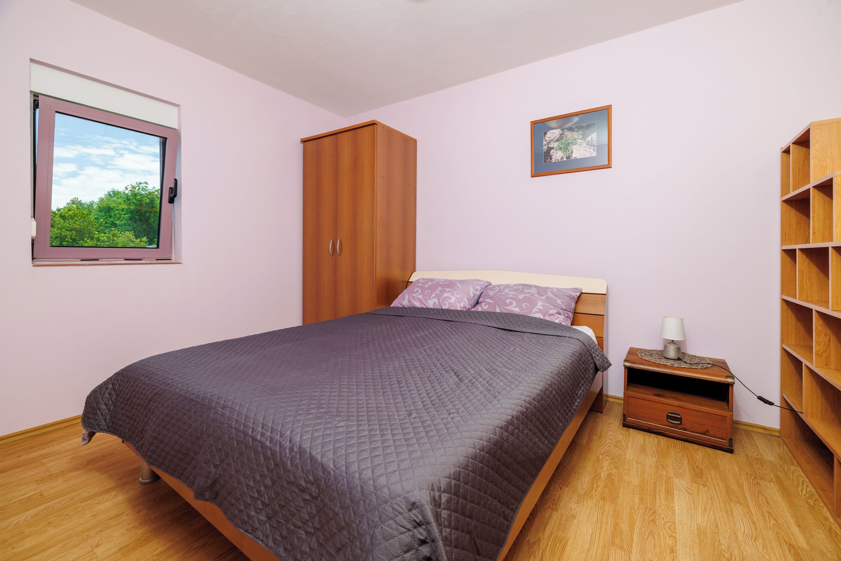 A bedroom with a bed, a wardrobe, a nightstand, a window, and a picture on the wall.