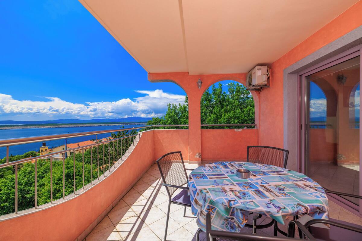 Un balcon avec une table et des chaises, donnant sur une étendue d’eau et sur de la verdure.