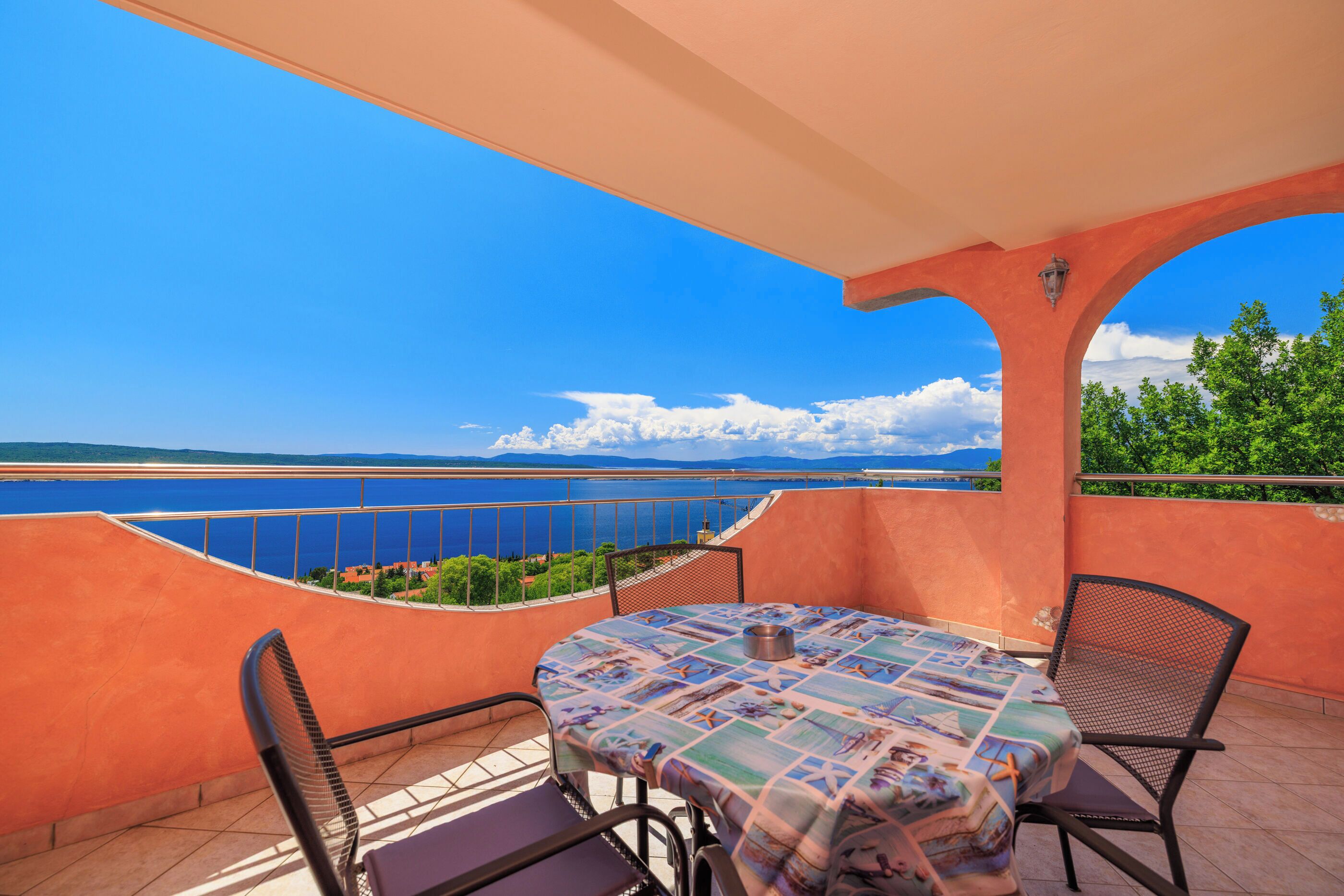 Un balcon avec une table et des chaises, donnant sur une étendue d’eau et sur une terre lointaine.