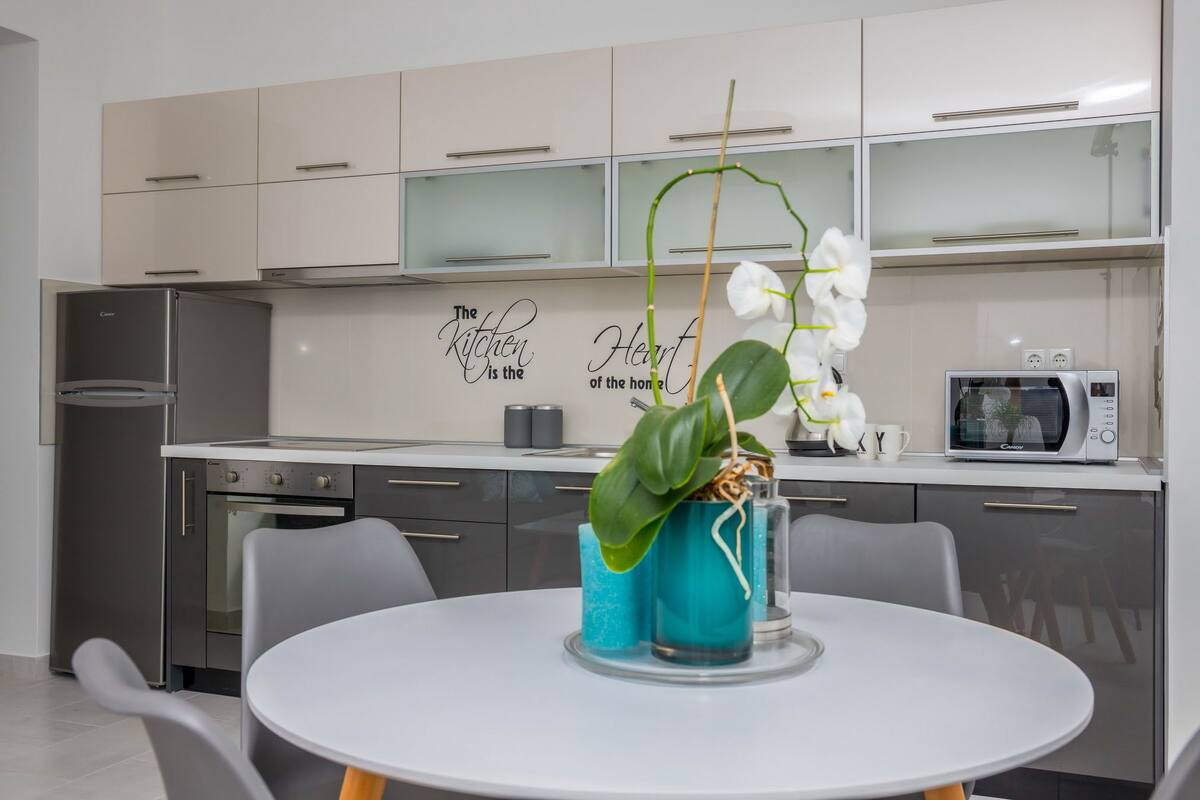 A modern kitchen with a white refrigerator, a microwave, and a sink. There is a table with chairs and a decorative orchid plant. The kitchen cabinets are light-colored with glass doors.