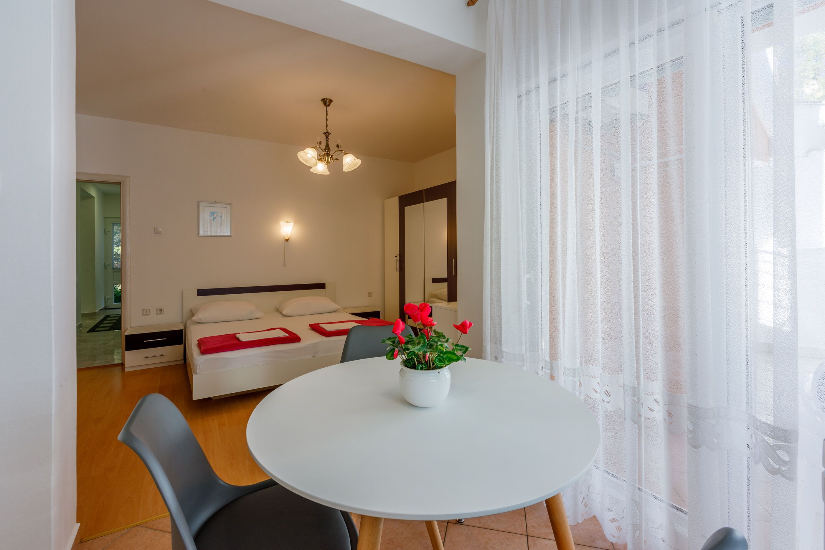 A modern hotel room with a round dining table, a white sofa, a bed with a red blanket, and a large window with sheer curtains.
