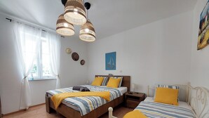A bedroom with two beds, wooden flooring, a window with sheer curtains, and a wall-mounted light fixture.