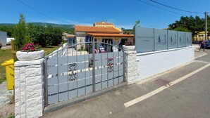 A gated entrance with a stone wall and metal gate, leading to a building with a tiled roof and a sign in a foreign language.