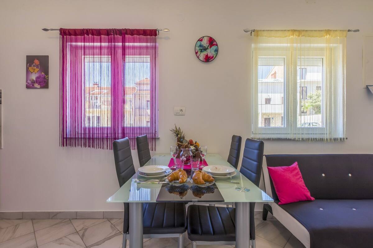 A dining area with a glass table, black chairs, and a black sofa with a pink pillow.