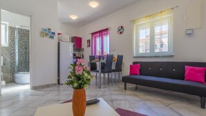 A modern living room with a black sofa, a white coffee table, and a vase of flowers. There is a dining area with chairs and a table, and a kitchen in the background. - Apartment Lena (Crikvenica)