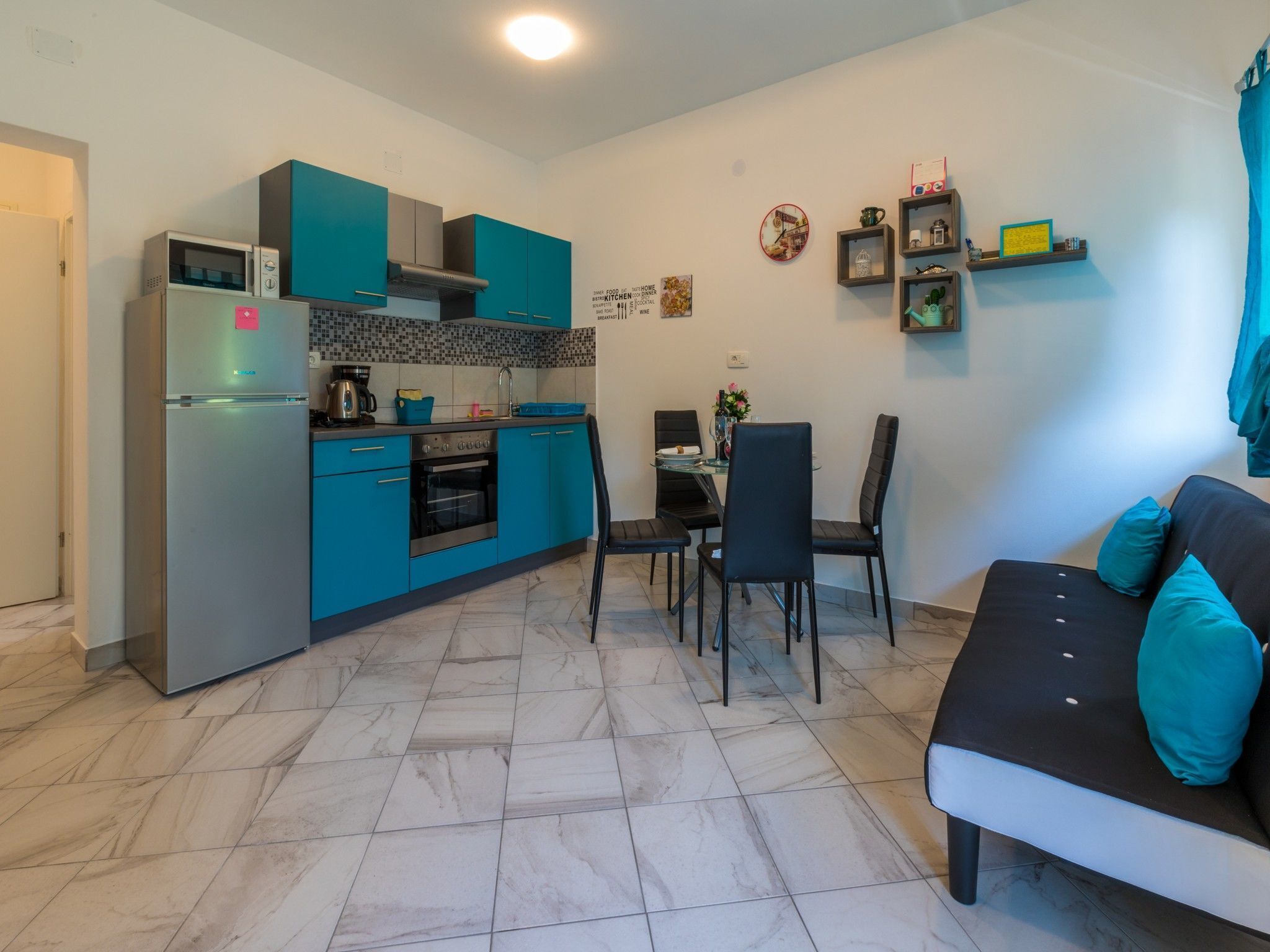 Une cuisine compacte avec des meubles de couleur turquoise, un réfrigérateur et une cuisinière. On y trouve un coin repas avec une table et des chaises, ainsi qu’un canapé agrémenté de coussins turquoise.