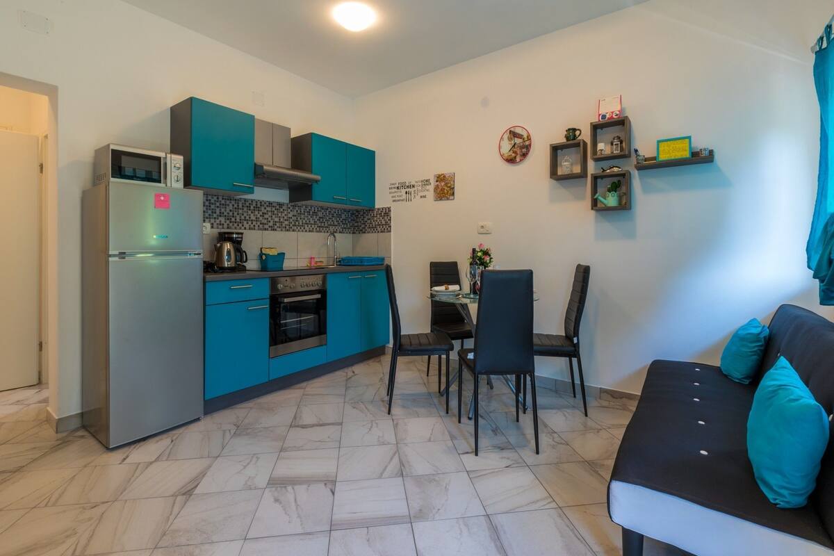 A compact kitchen with teal cabinets, a refrigerator, and a stove. There is a dining area with a table and chairs, and a sofa with teal cushions.