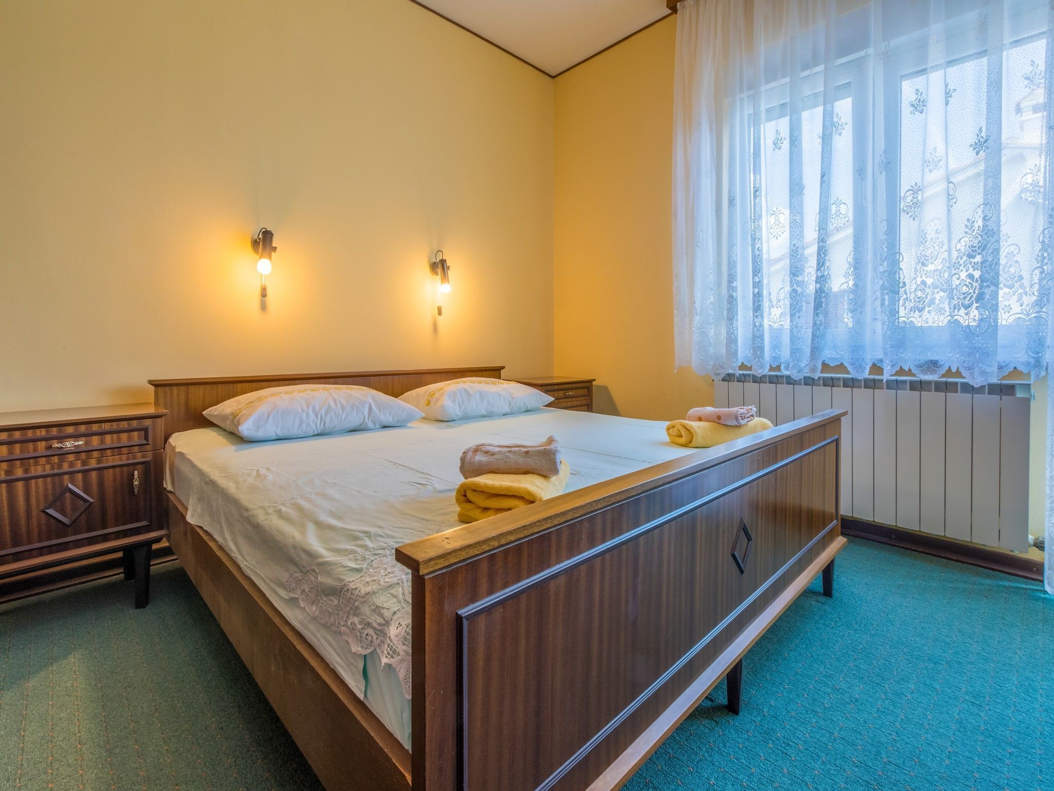 Una camera da letto con un letto in legno, due lampade a parete, una finestra con tende in pizzo e un termosifone.