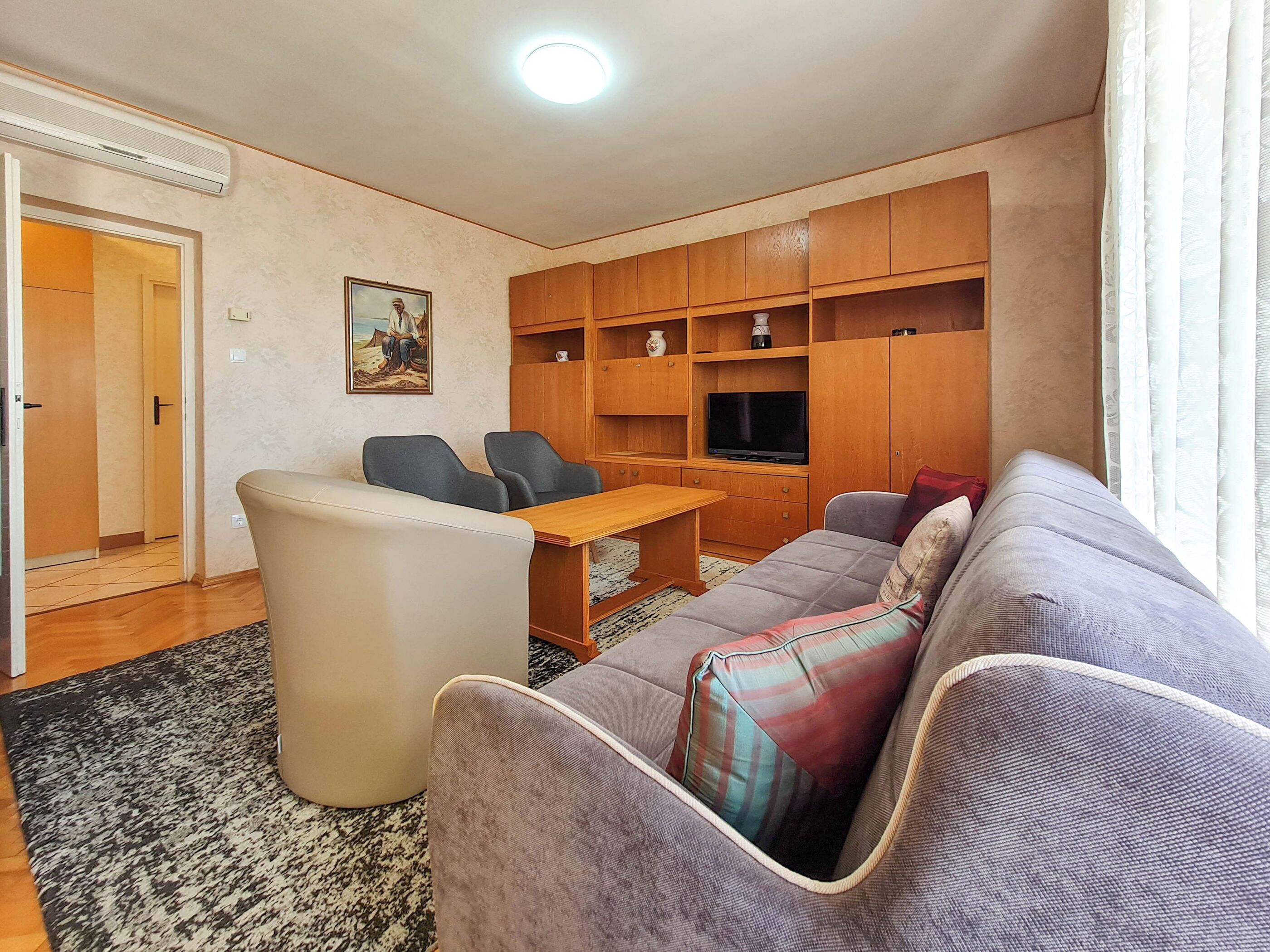 Een woonkamer met een bank, twee fauteuils, een salontafel en een televisie. Er is een houten kast met planken, een schilderij aan de muur en een deur die naar een andere kamer leidt.
