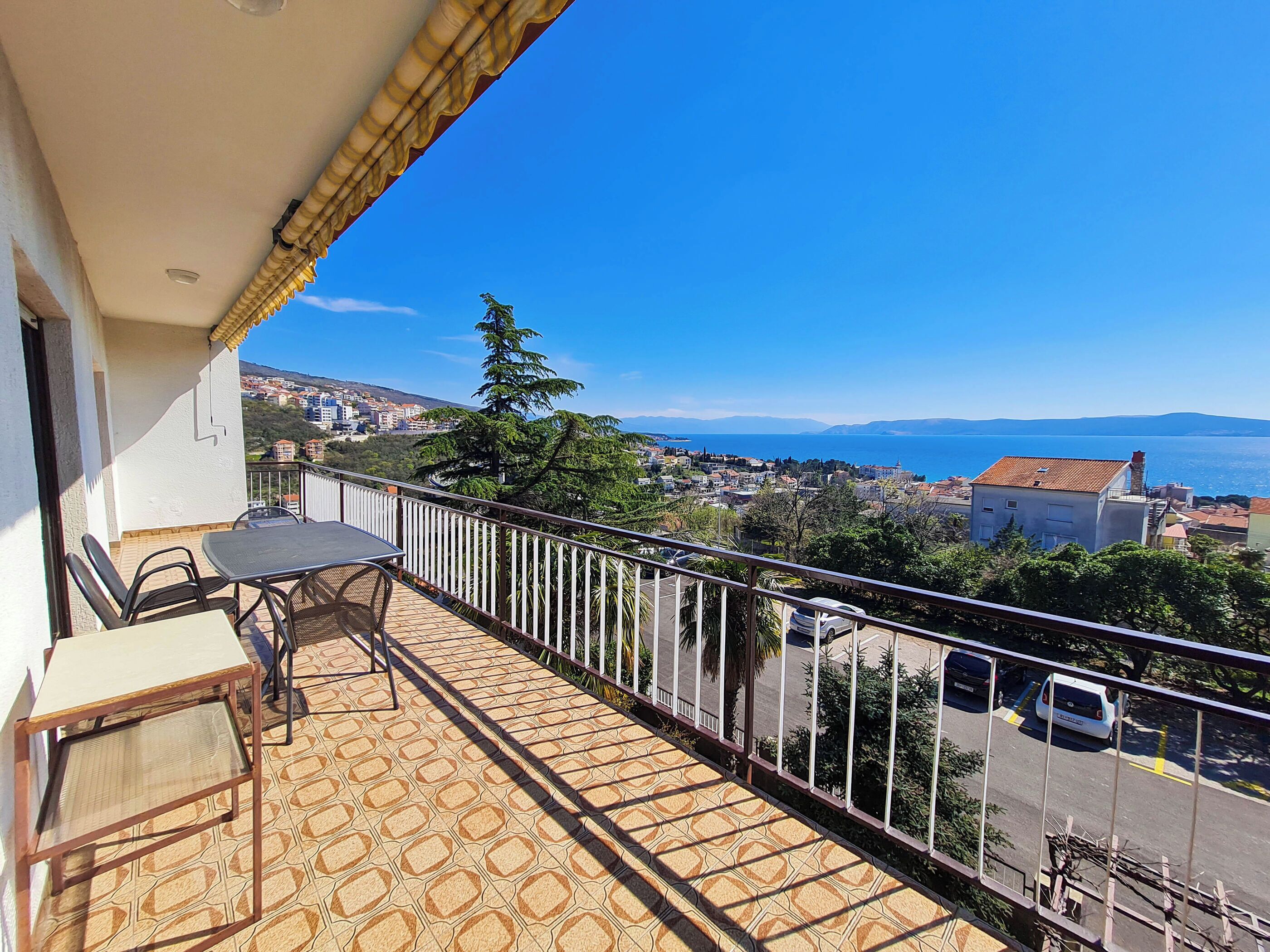 A balcony with a table and chairs, overlooking a coastal town and the sea.