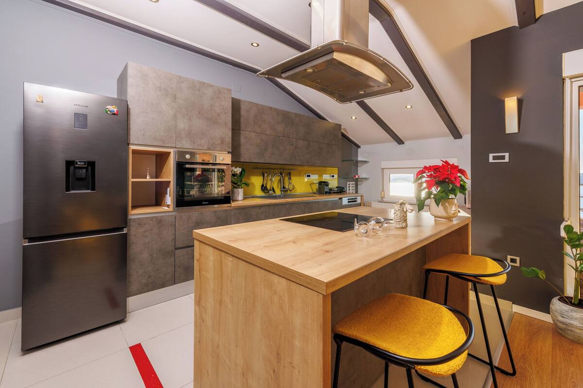 A modern kitchen with a stainless steel refrigerator, a wooden island with two yellow bar stools, a microwave, and a built-in oven.