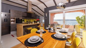 Une cuisine moderne avec un coin repas, comprenant une table en bois dressée pour un repas, une grande fenêtre donnant sur la mer et un four encastré.