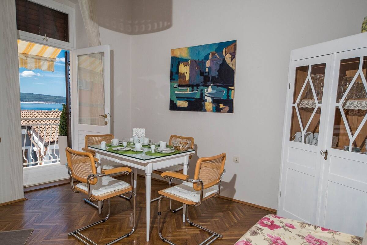 A dining area with a glass table, wicker chairs, and a view of the sea through an open door.