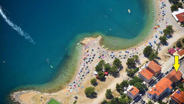 An aerial view of a beach with clear waters, a sandy shore, and a row of buildings with red roofs. - Apartments Latino (Crikvenica)