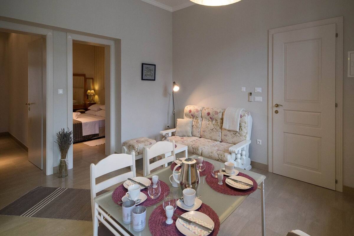 A cozy living room with a sofa, a dining table set for two, and a door leading to another room.