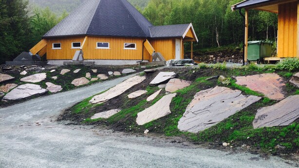 Una cabaña de madera con techo empinado, rodeada de un paisaje rocoso y un bosque denso.