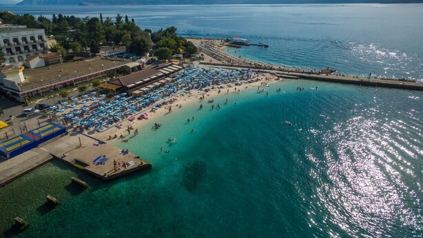 Ein Luftbild zeigt einen Strand mit klarem türkisblauem Wasser, einen Steg und eine Reihe von Strandsonnenschirmen.
