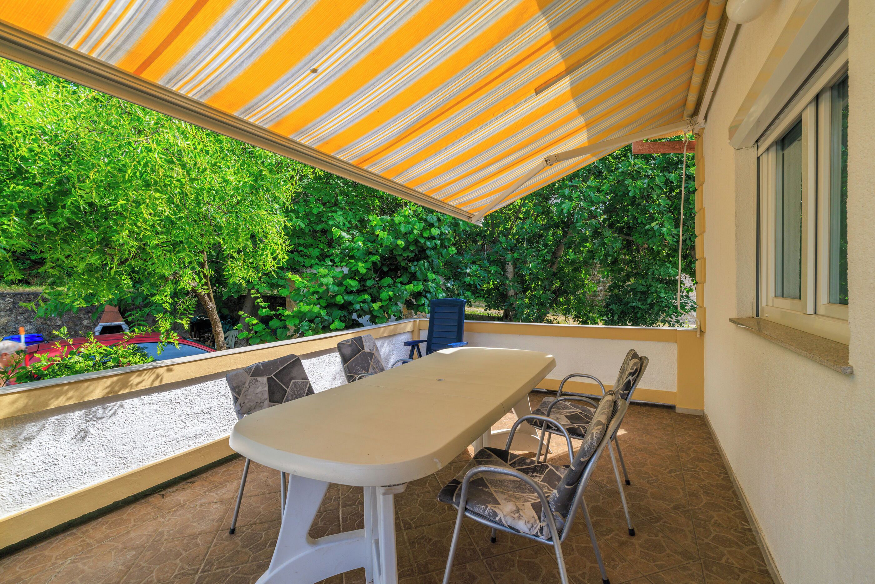 Une terrasse extérieure couverte, avec une table et des chaises, entourée de verdure et offrant une vue sur la rue.