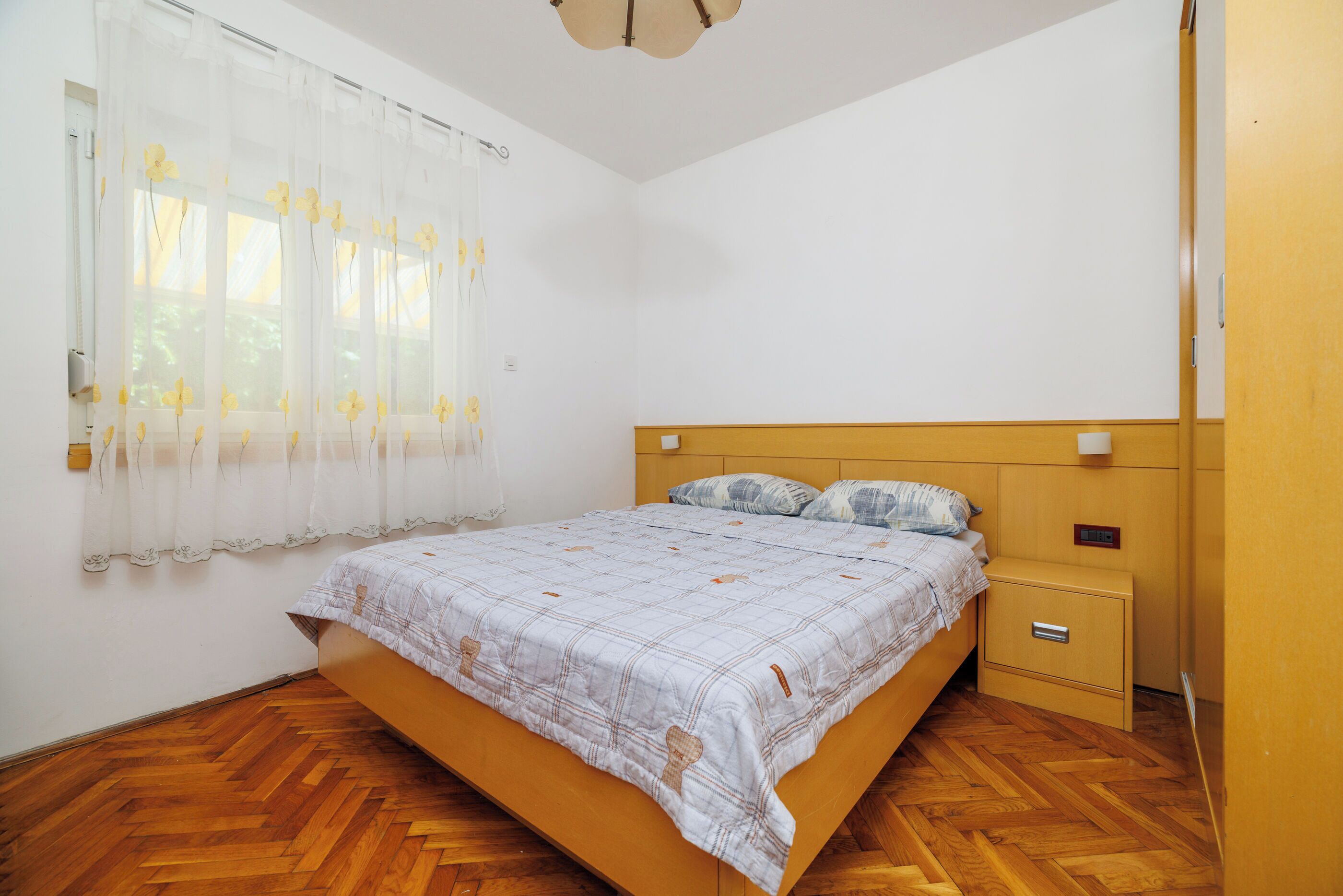 A bedroom with a wooden floor, a bed with a patterned bedspread, a nightstand, and a window with sheer curtains.