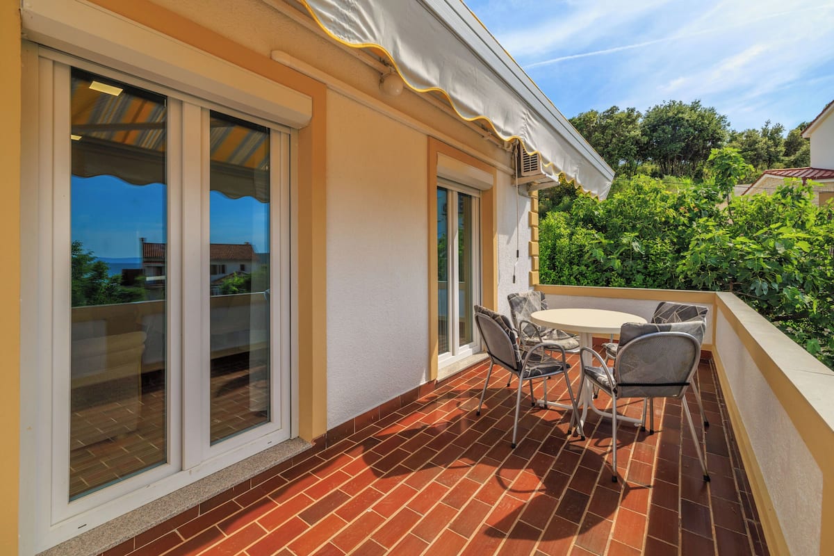 A patio with a table and chairs, a sliding glass door, and a view of greenery and buildings.