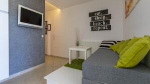 A modern living room with a grey sofa, a white coffee table, and a flat-screen TV mounted on a grey wall. There is a small white side table with a vase and a framed picture. A motivational quote is displayed on the wall. - Apartment Color Green (Dramalj)