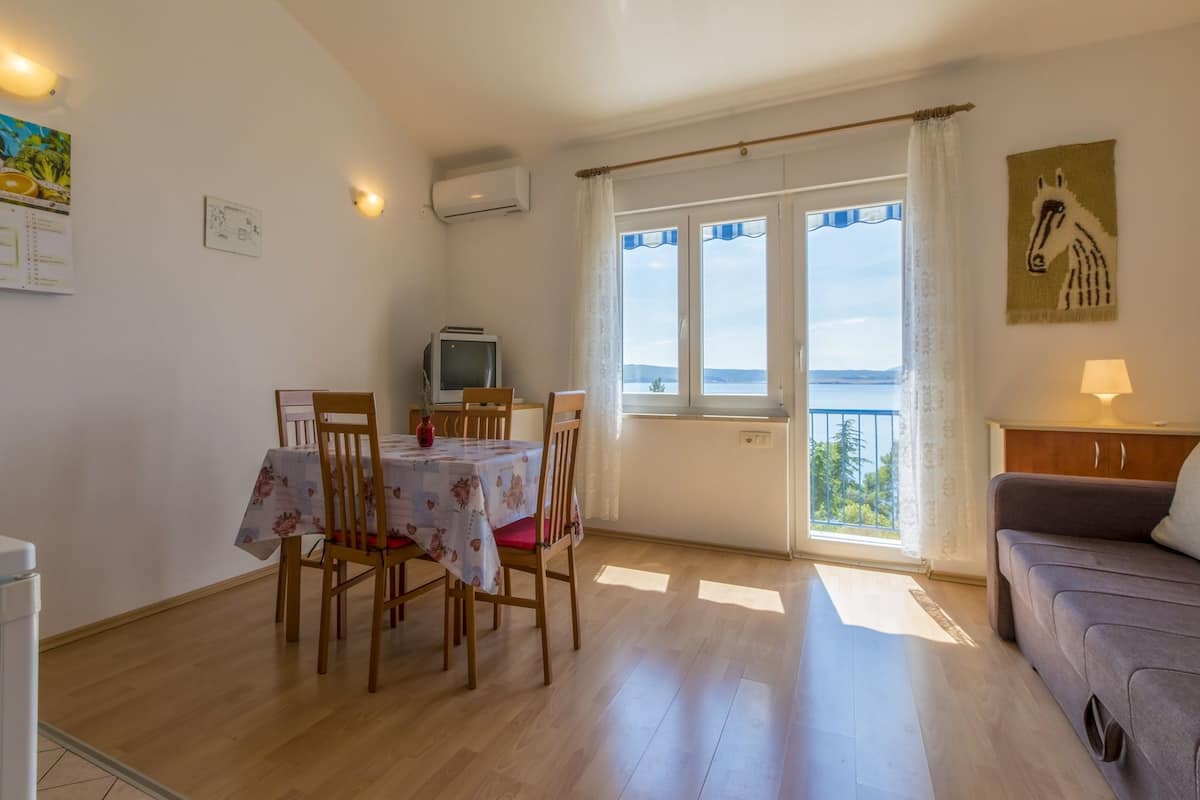 A dining area with a table set for four, wooden chairs, a small television on a stand, and a view of the sea through large windows.
