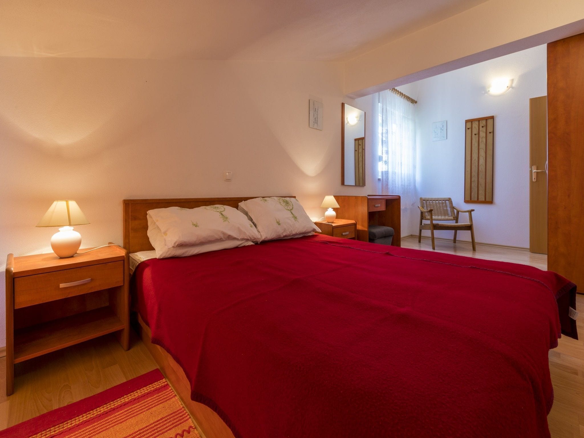 Une chambre à coucher avec un lit en bois, deux tables de chevet avec des lampes, une couverture rouge, une chaise et un miroir au mur.