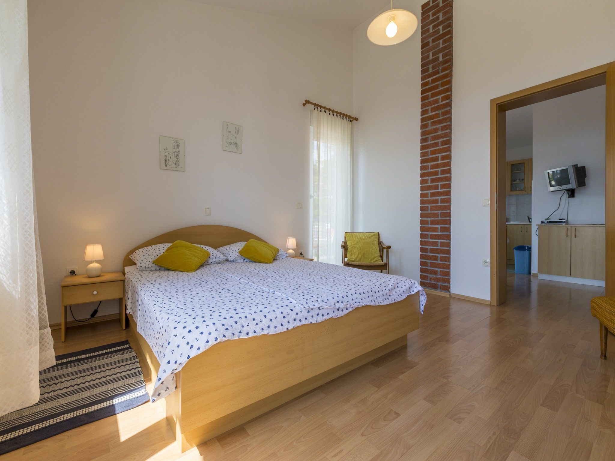 Ein Schlafzimmer mit einem Holzbett, einem Nachttisch, einem Sessel, einem Fernseher und einer sichtbaren Küchenzeile durch eine geöffnete Tür.