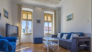 A living room with a blue sofa, a white coffee table, and a TV on a stand. There are two windows with curtains, and a painting on the wall. - Apartment Piano (Crikvenica)
