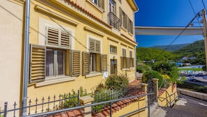 A yellow building with multiple windows and shutters, a balcony, and a view of a green landscape and a road with vehicles. - Apartment Piano (Crikvenica)