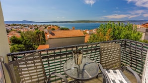 Ein Balkon mit Tisch und Stühlen, der auf eine Küstenstadt mit roten Dächern und blauem Himmel blickt.