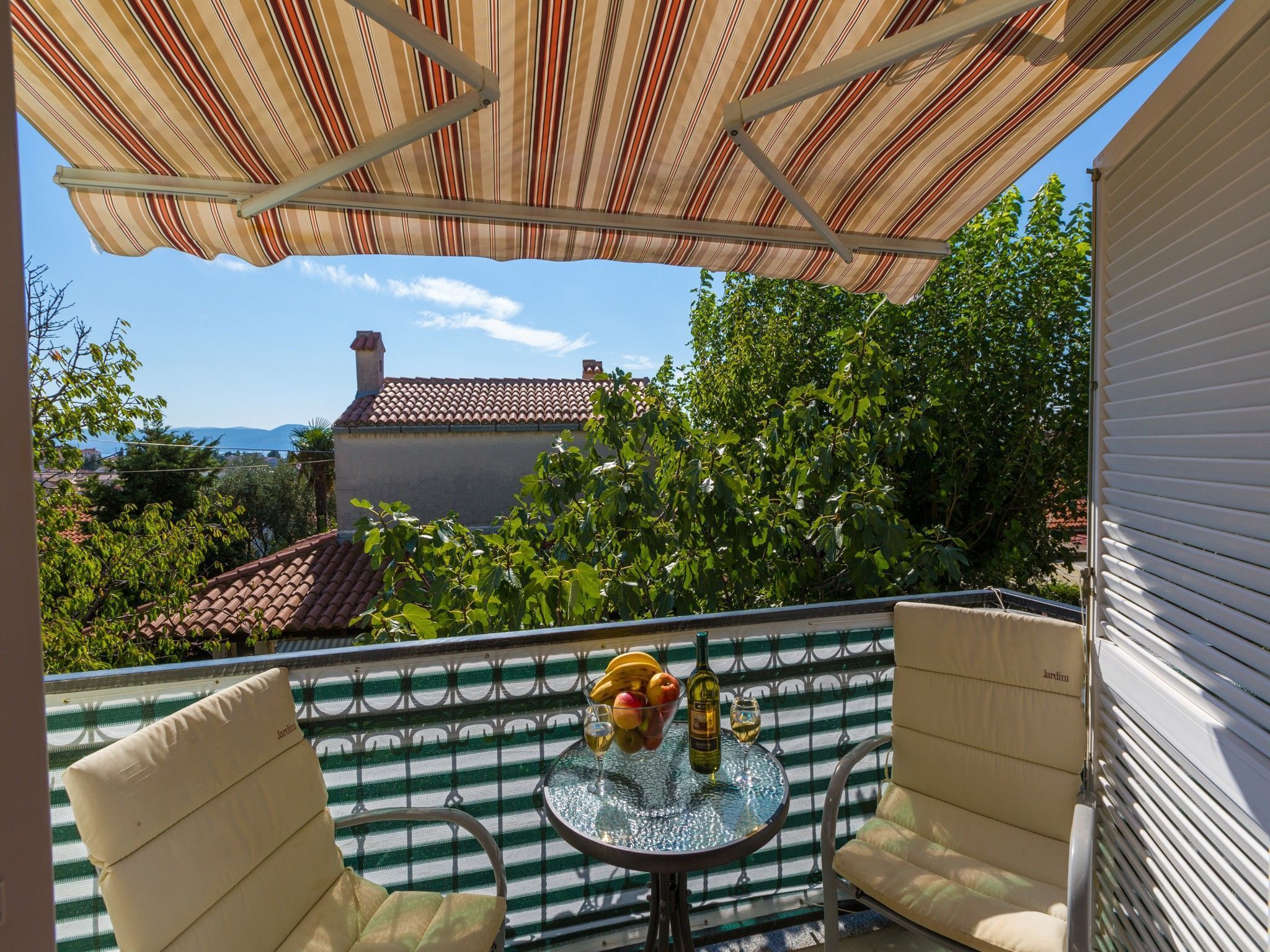 A balcony with a striped awning, two cushioned chairs, a small round table with a bottle and fruit, and a view of a house with a tiled roof and greenery.