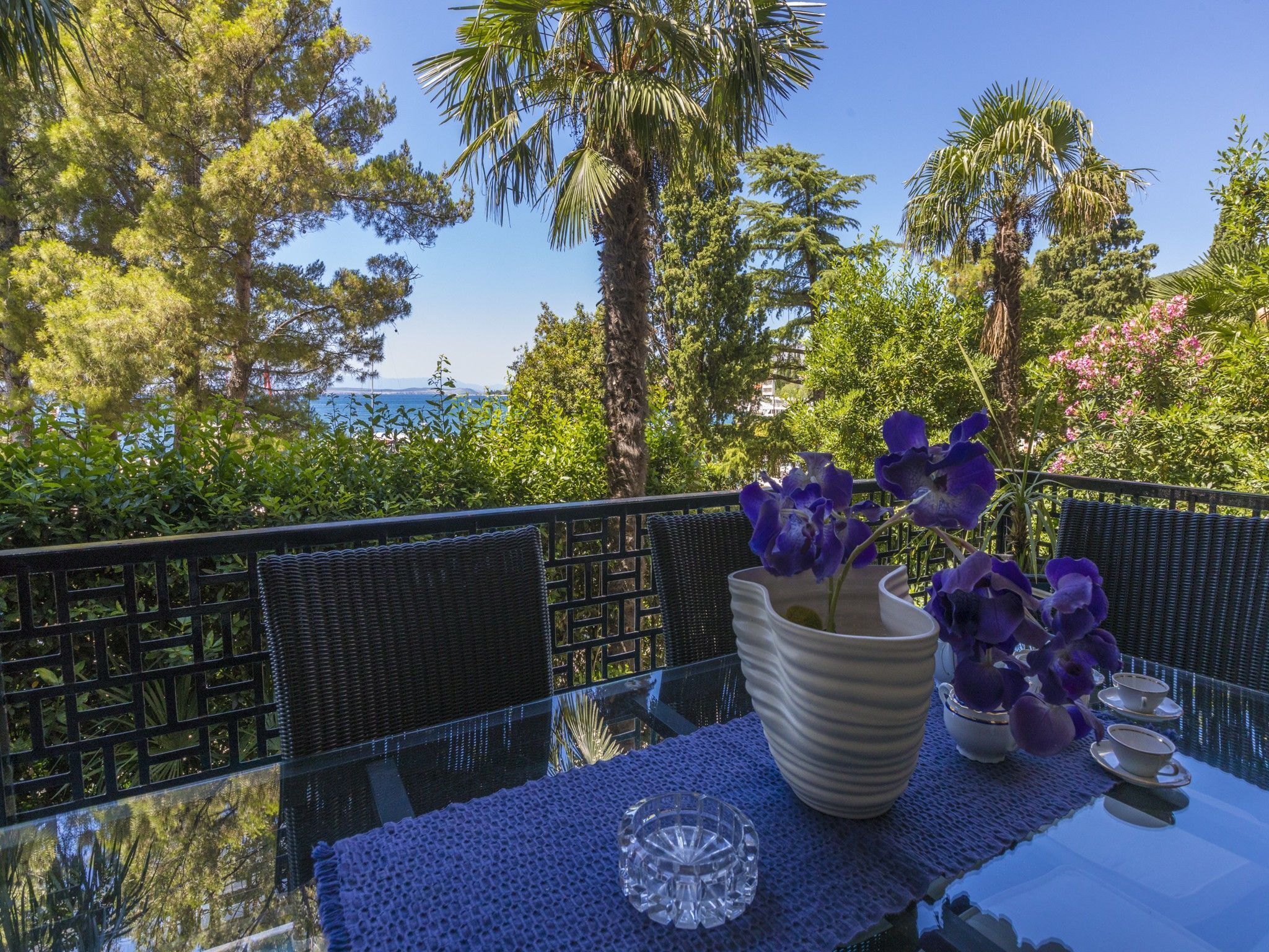A balcony with a glass table, a white vase with purple flowers, and a clear glass bowl.