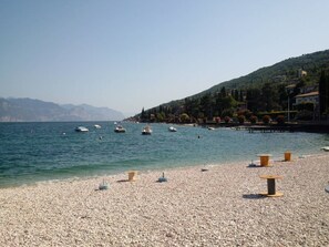 Ein Kiesstrand mit Booten auf dem Wasser und Häusern im Hintergrund.