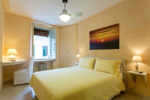 Ein Hotelzimmer mit einem Bett, zwei Nachttischen mit Lampen, einem Fenster mit Rollläden, einem Heizkörper und einem Sonnenuntergangsbild an der Wand.
