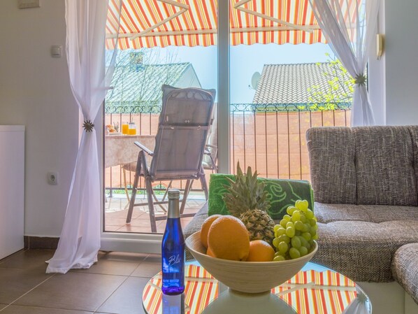 A room with a glass table holding a fruit bowl, a blue bottle, and a striped tablecloth. There is a wicker chair and a sofa with a cushion.