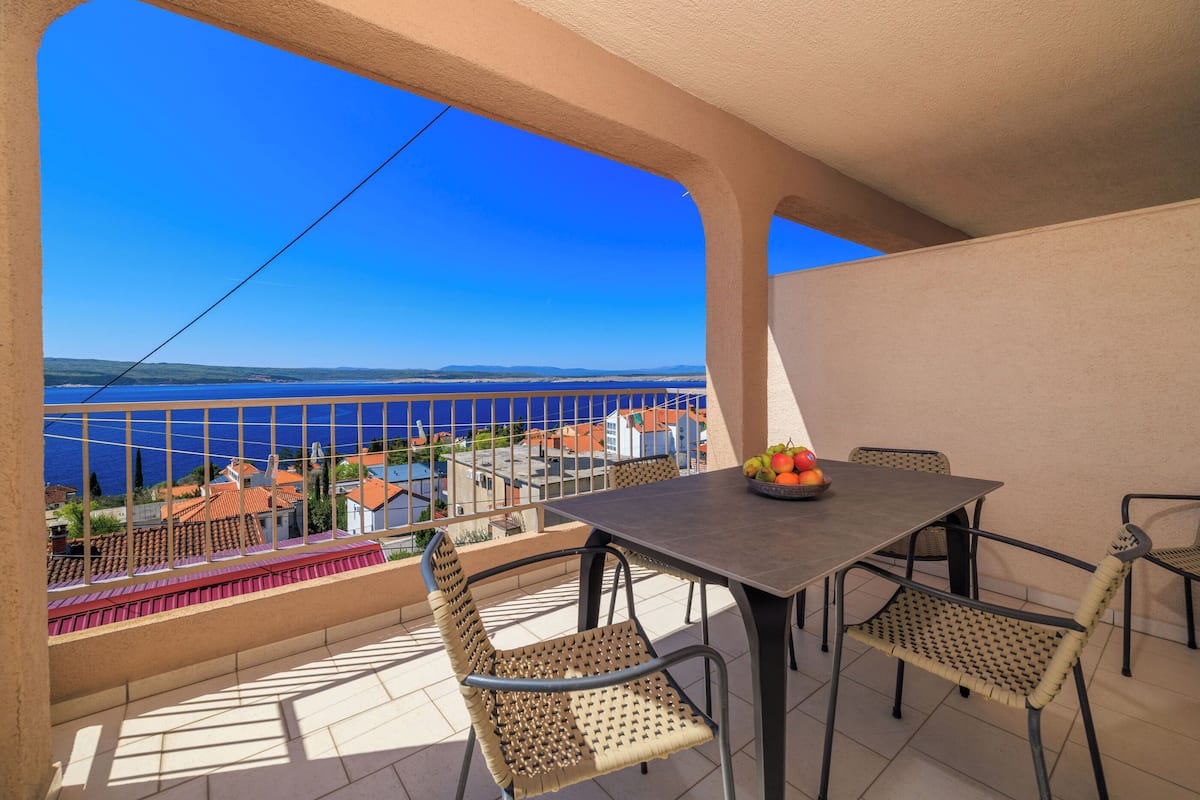 Ein Balkon mit Tisch und Stühlen, einer Obstschale und Blick auf das Meer und die Gebäude.