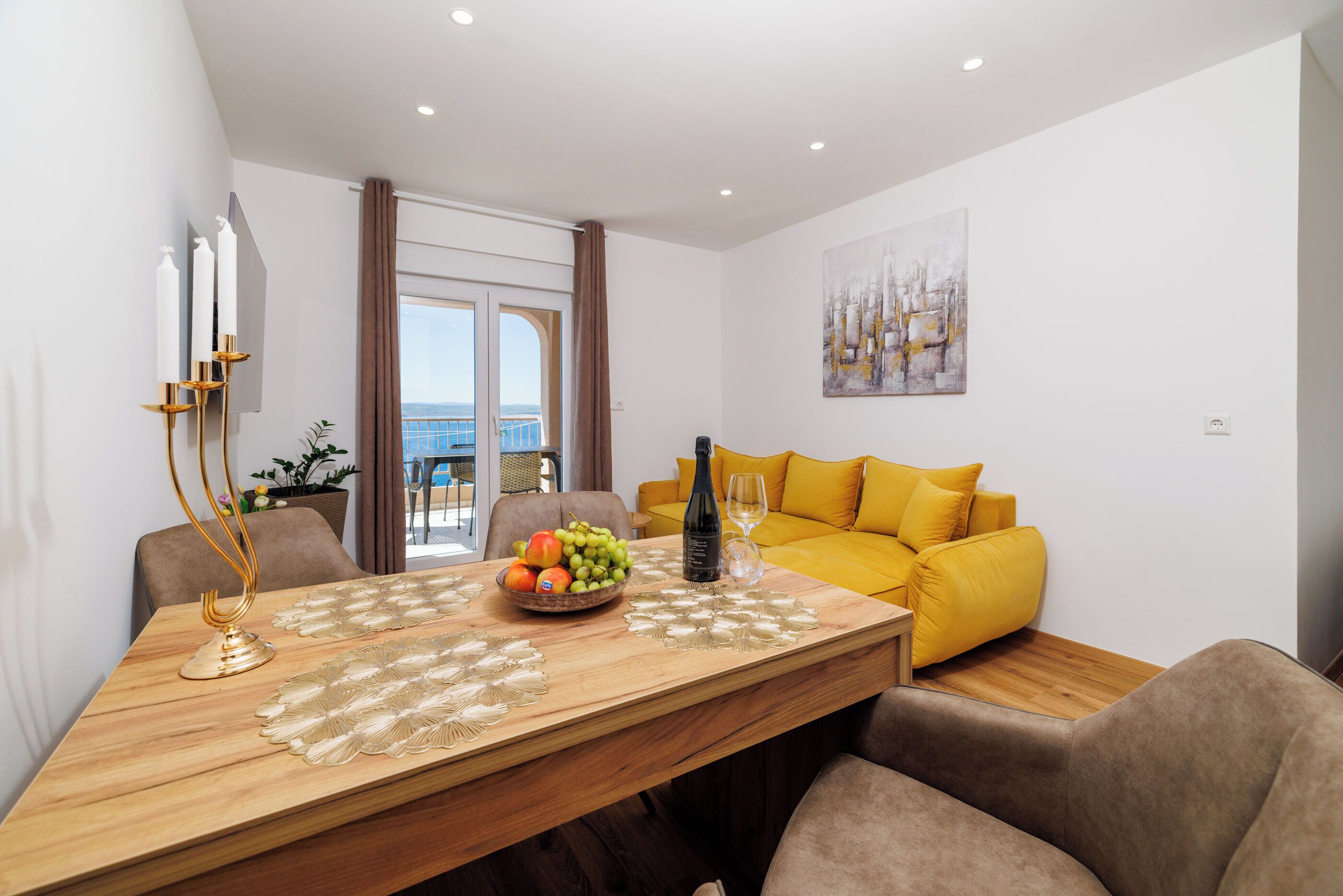 Een eetruimte met een houten tafel, stoelen, een schaal fruit, een wijnfles en een gele bank.