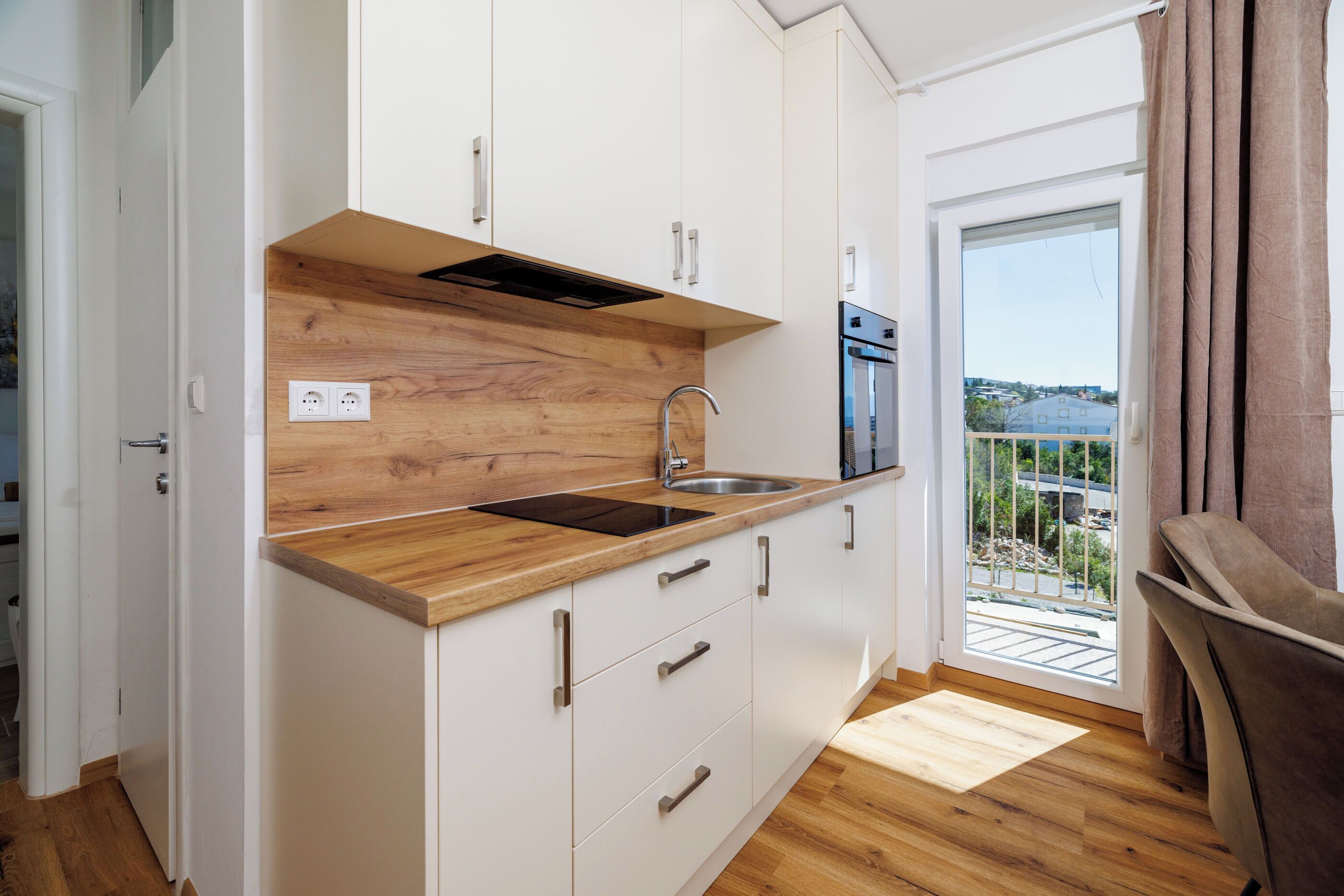 A modern kitchen with white cabinets, a wooden countertop, a stainless steel sink, and a view of the outside through a sliding door.