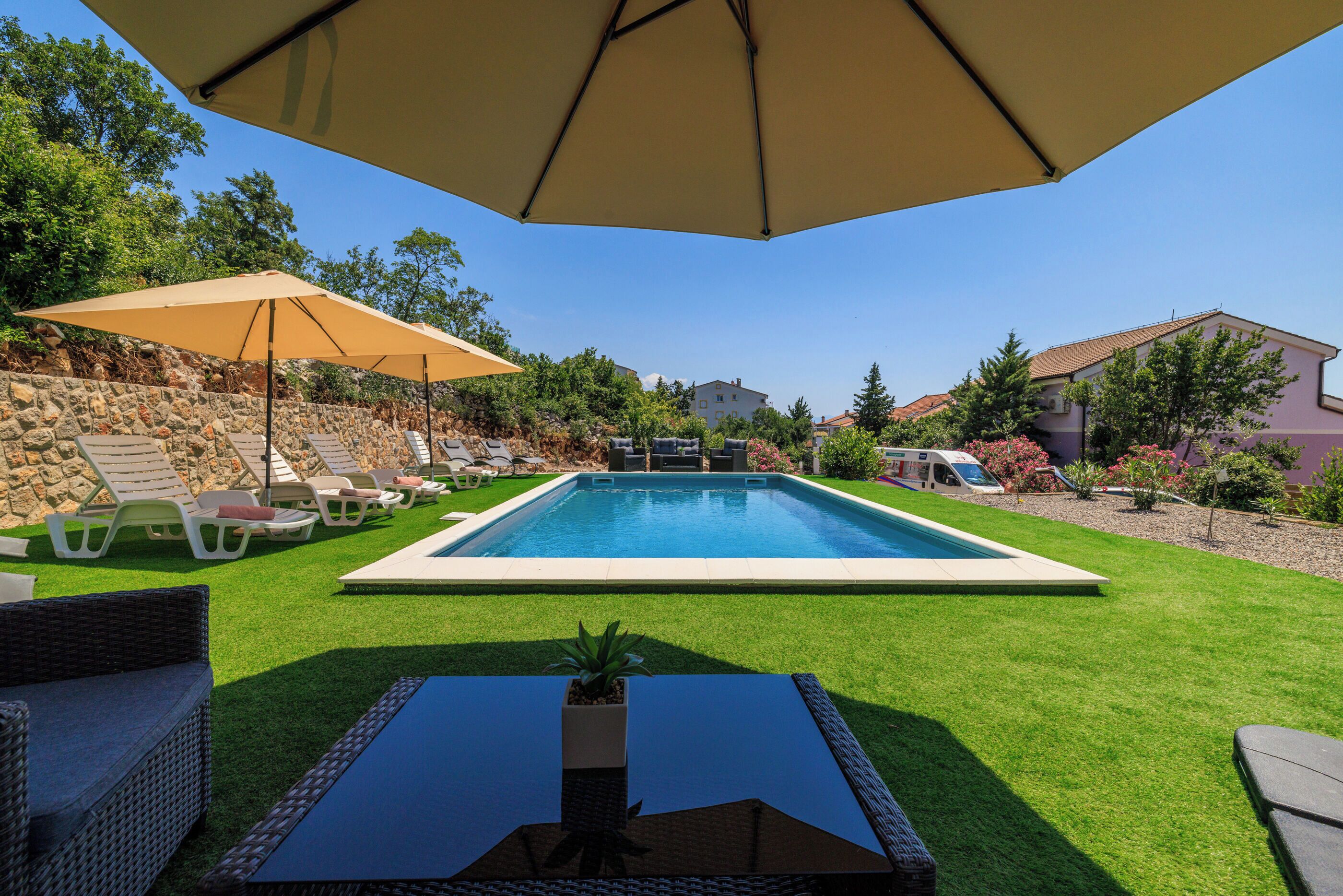 Une piscine rectangulaire entourée d’une pelouse, avec des chaises longues et une table agrémentée d’une plante.