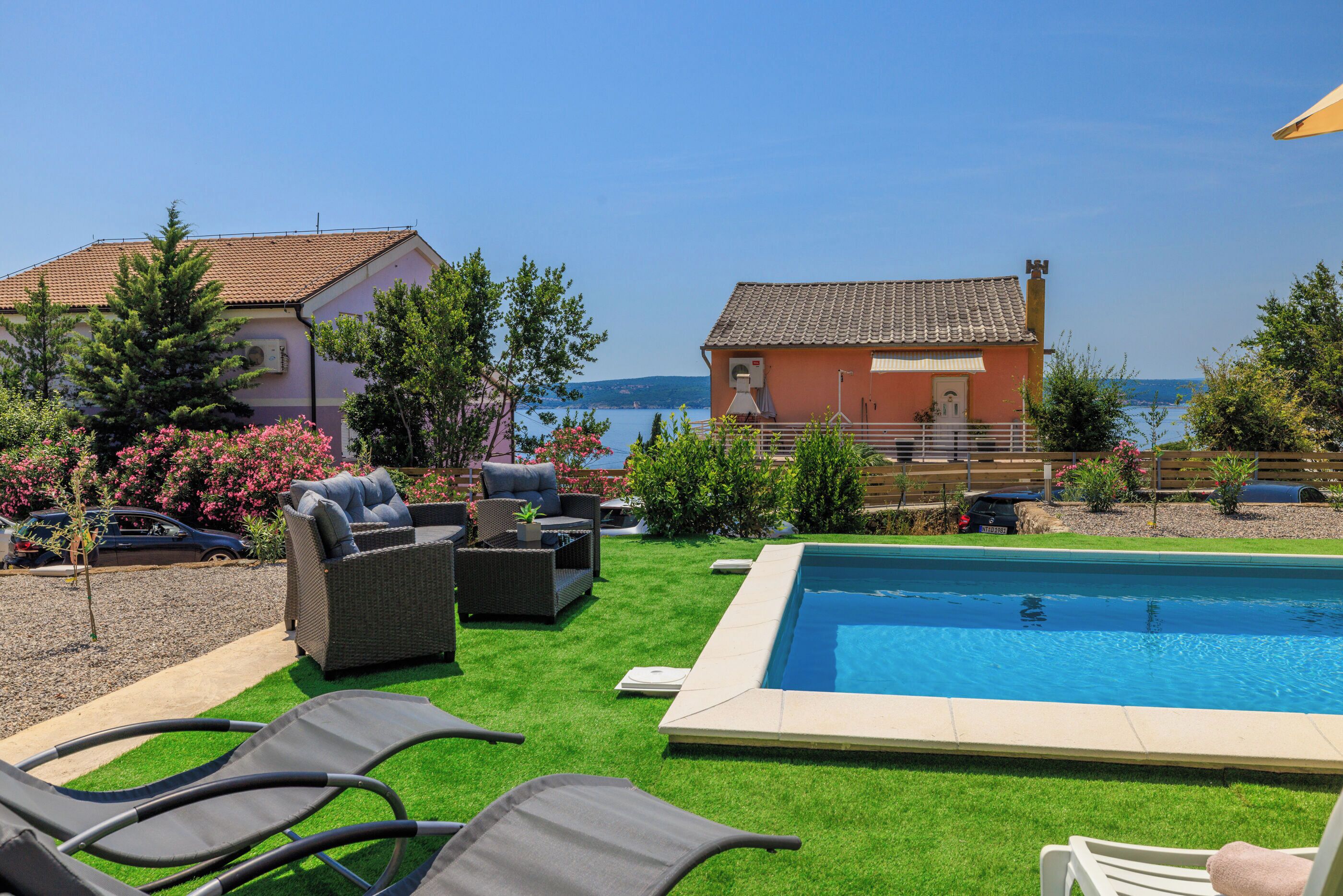 A pool area with a view of the sea, surrounded by greenery and outdoor furniture.