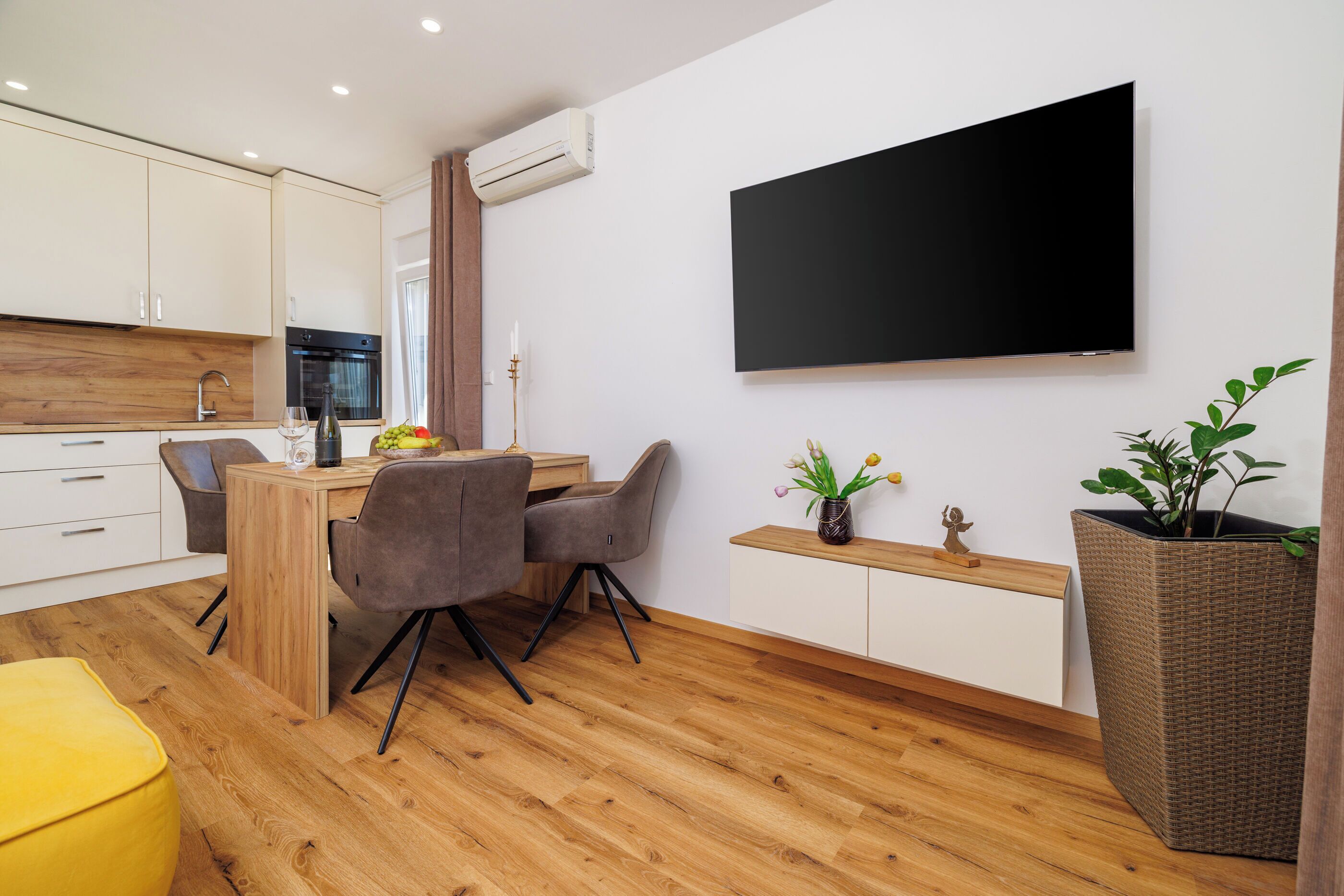 Una cucina moderna con mobili in legno, forno integrato, zona pranzo con tavolo e sedie in legno, TV a schermo piatto montata a parete e una pianta in vaso.