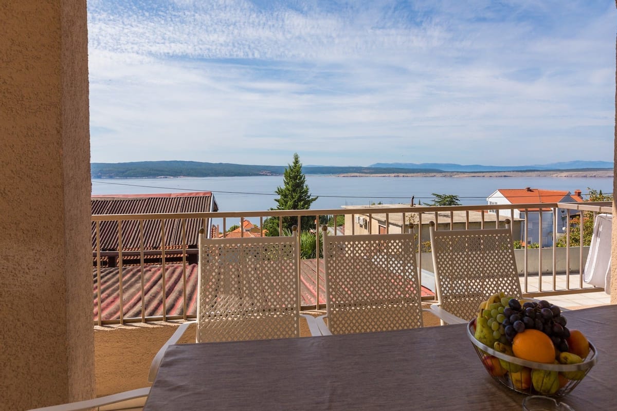 Ein Balkon mit Tisch, Stühlen und einer Obstschale. Durch die Brüstung ist eine malerische Aussicht auf ein Gewässer und ferne Berge sichtbar.