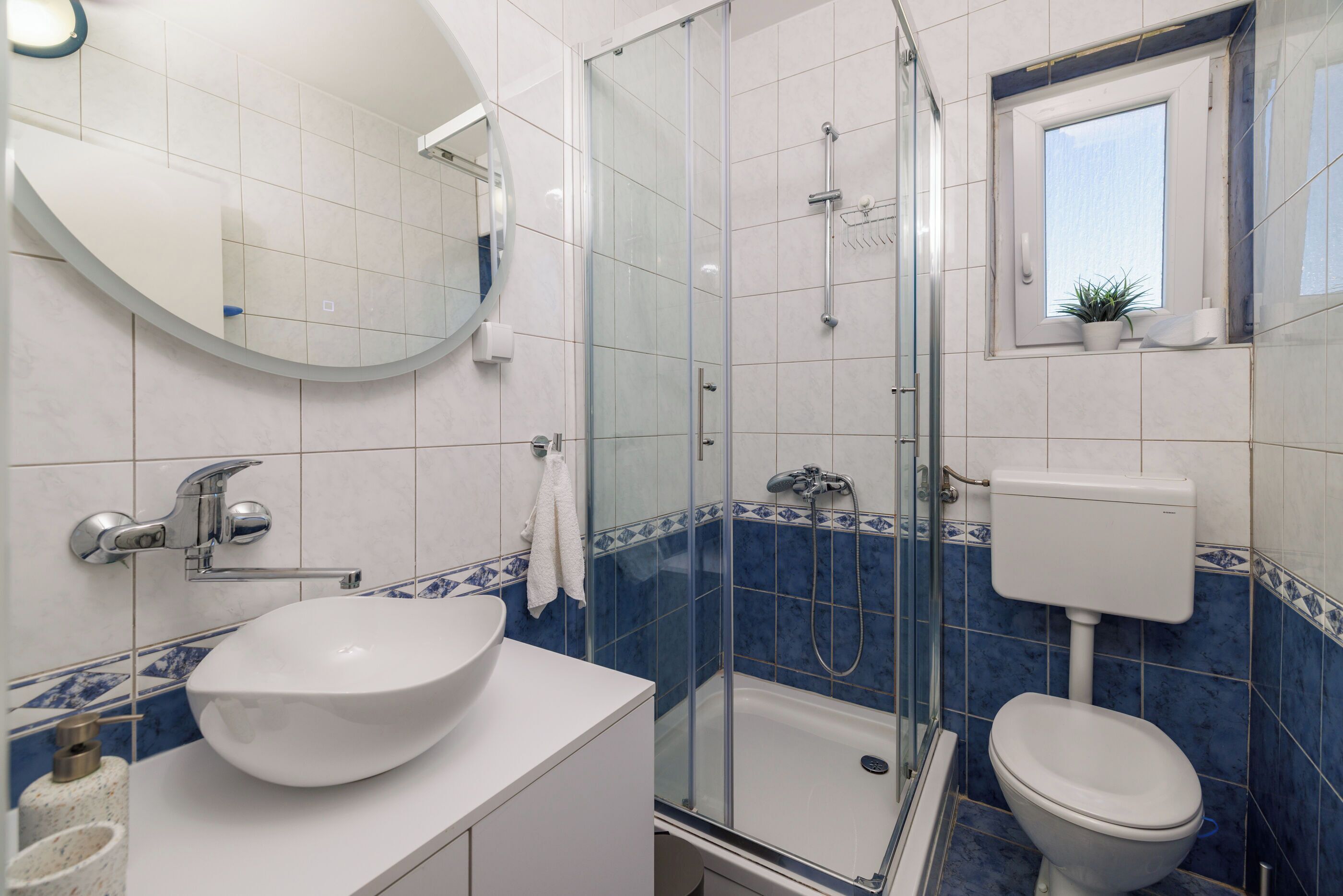 Un baño con lavamanos blanco, ducha con mampara de vidrio, inodoro y una pequeña ventana con una planta.