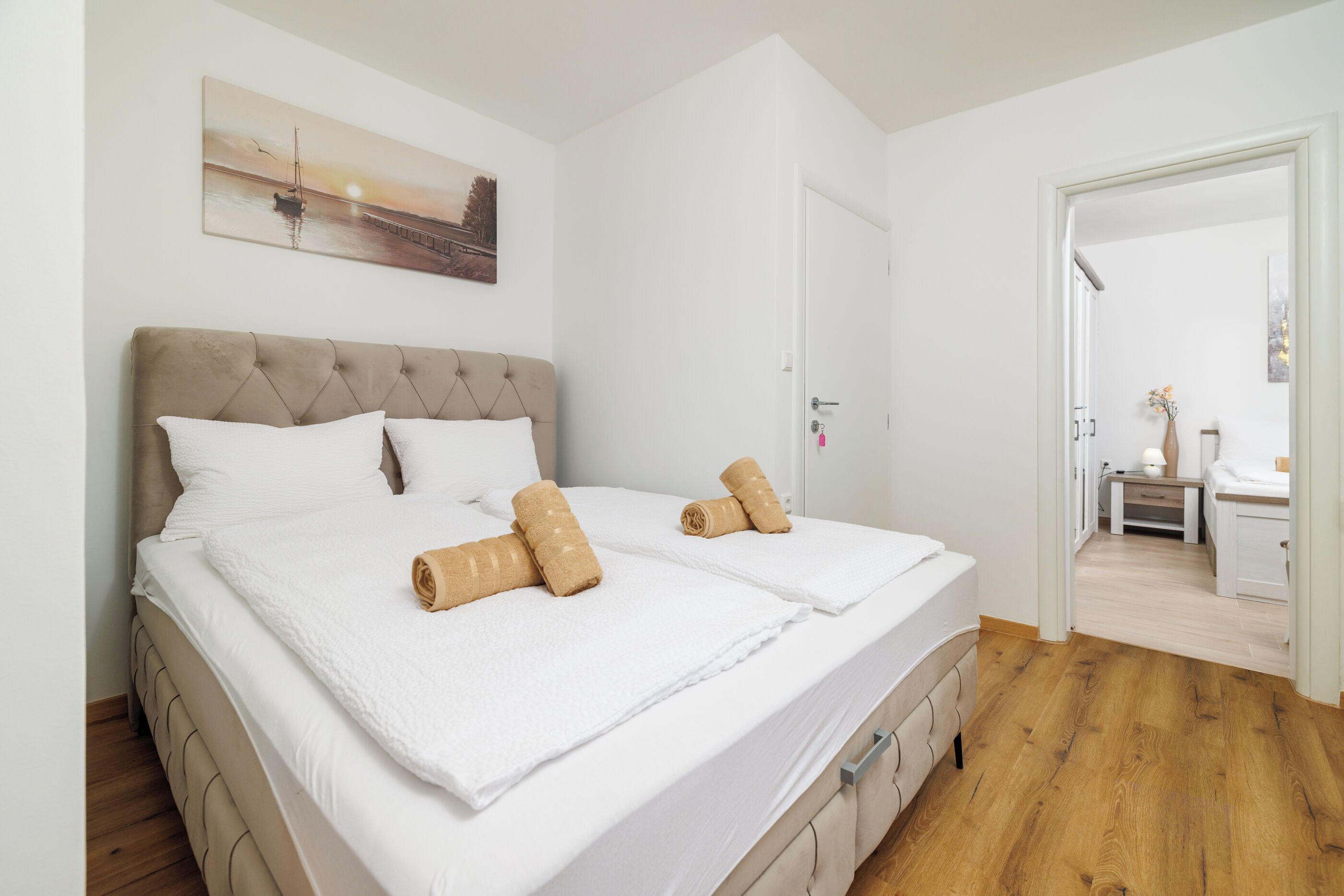 A double bed with a headboard, white bedding, and rolled towels. There is a painting above the bed, a door to the right, and a hallway leading to another room.