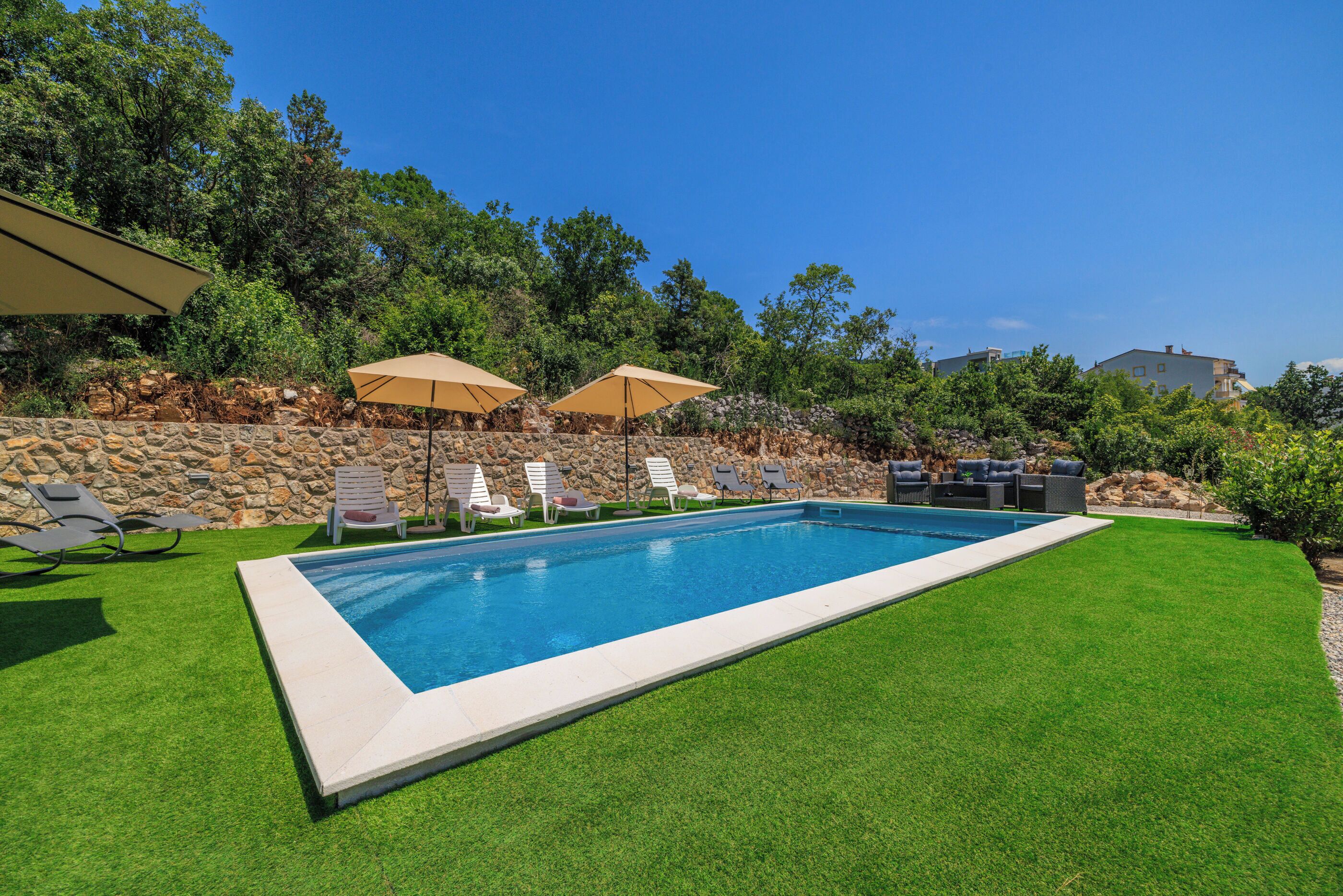A rectangular swimming pool with a stone wall and green lawn. There are lounge chairs and umbrellas.