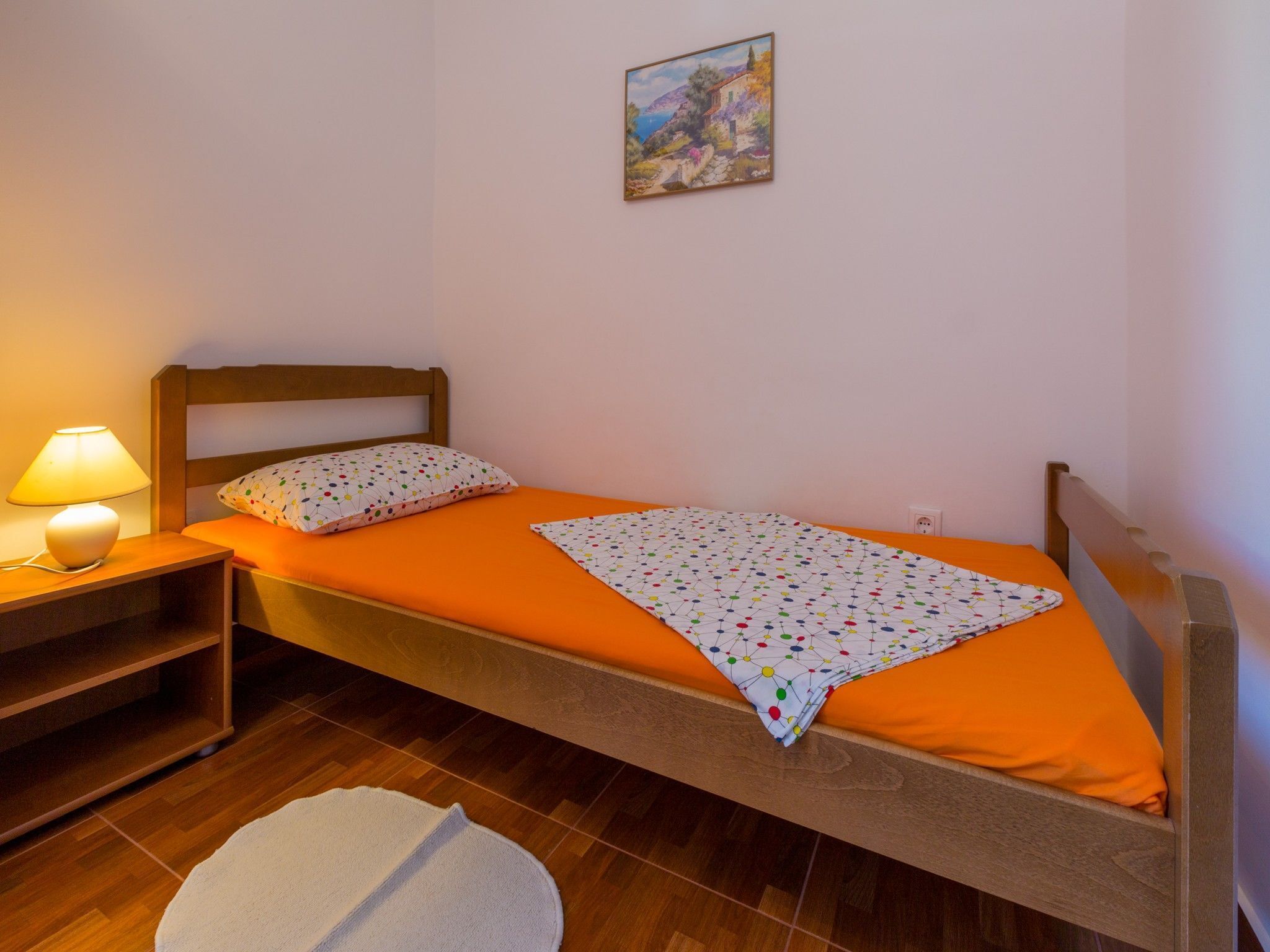 A single bed with an orange bedspread, a white pillow with a colorful pattern, and a wooden headboard. There is a small bedside table with a lamp and a white rug on the floor.