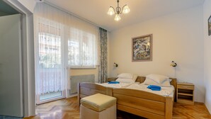 A bedroom with a wooden bed, a window with sheer curtains, a radiator, a nightstand, and a wall-mounted lamp. - Apartments Villa 7 (Crikvenica)